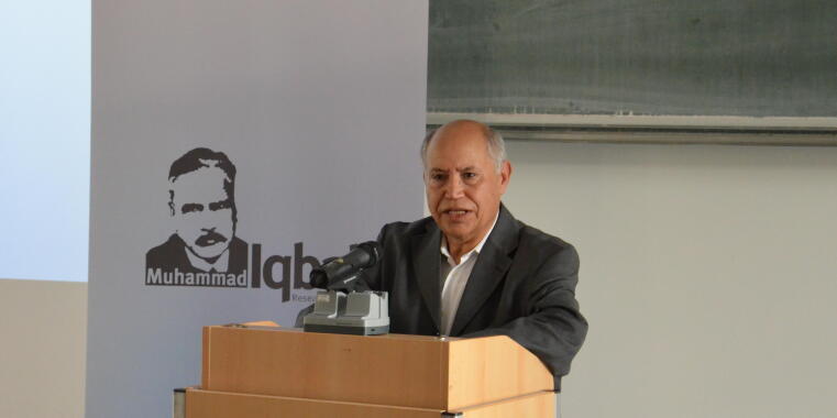 Prof. Dr. Mohamed Turki hinter einem Rednerpult vor einem Plakat mit dem Konterfei Muhammad Iqbals
