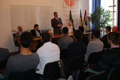 Prof. Dr. Halil ef. Mehtić stehend hinter einem Schreibtisch rechts von ihm zwei Männer, links eine Frau im Hintergrund drei Fahnen vor dem Schreibtisch sitzen Menschen auf Stühlen mit dem Rücken zum Betrachter