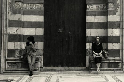 Zwei Frauen sitzen rechts und links neben einem Eingang zu einem großen Gebäude