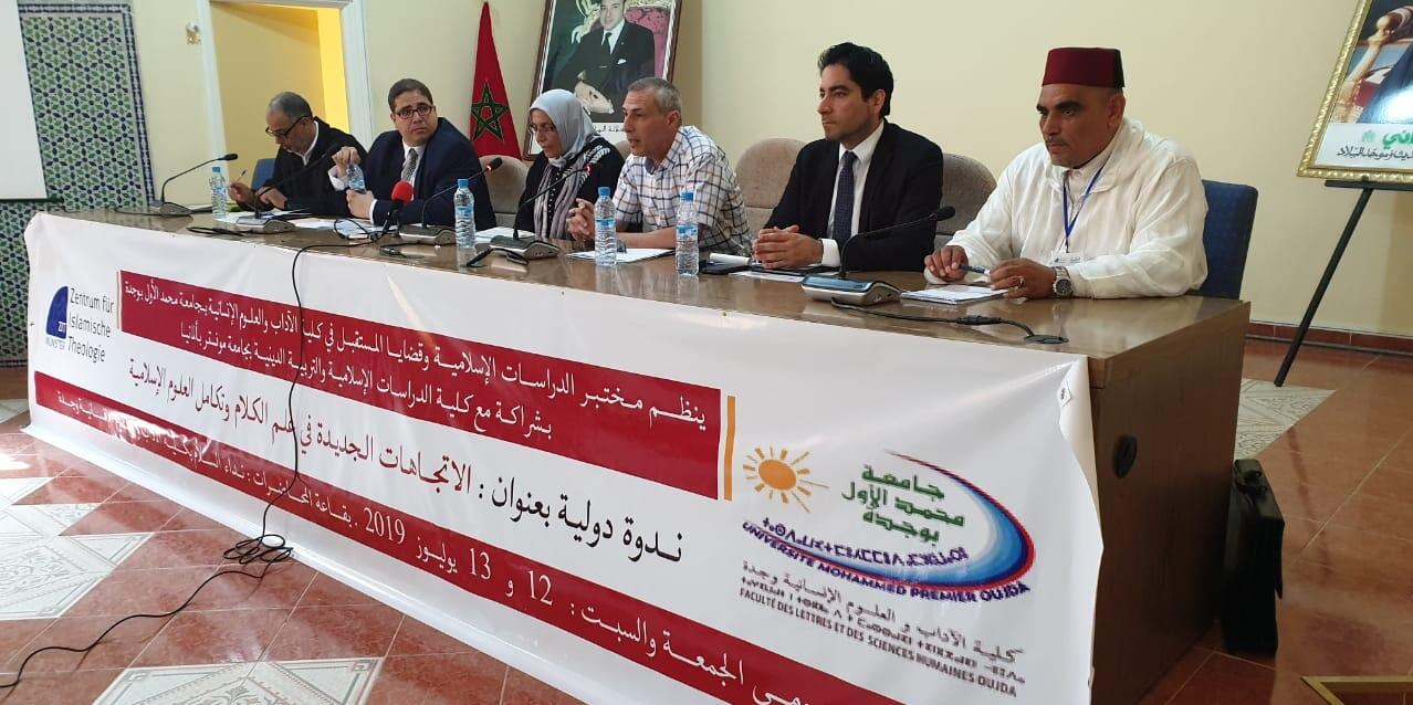 Hinter einem Tisch vor dem ein Plakat mit arabischer Schrift hängt sitzen sechs Personen