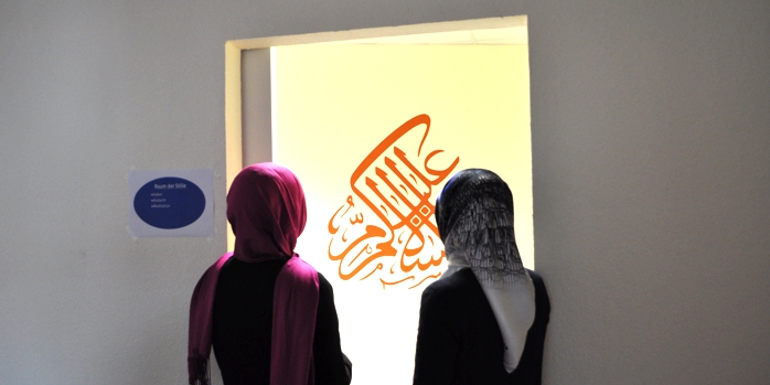 Zwei Menschen mit Kopftuch mit dem Rücken zum Betrachter stehen vor einer Kaligraphie