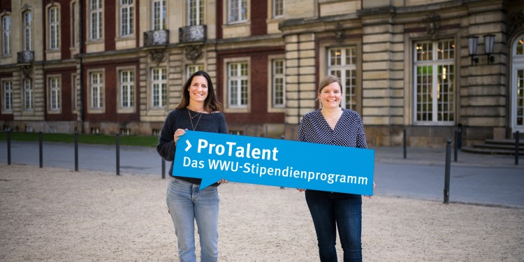 Christiane Borgmann und Sabrina Dirksen vom Team von ProTalent zeigen vor dem Schloss das Schild des WWU-Stipendienprogramms.<address>© WWU – Thomas Mohn</address>
