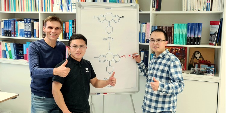Prof. Dr. Armido Studer mit Dr. Hui Cao und Dr. Qiang Cheng (v. l.). Auf der Tafel ist die chemische Transformation von Loratadin dargestellt, einem wichtigen Histamin-H1-Rezeptor-Antagonisten, welcher durch eine Trifluormethylgruppe modifiziert wurde.<address>© WWU - Arbeitskreis Studer</address>