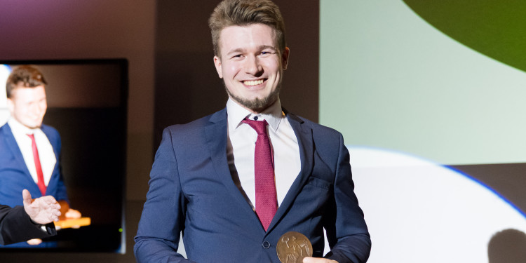 Der Preisträger Maurice Dellin hält die Universitätmedaille in den Händen und lacht in die Kamera.<address>&copy; UKM - Marcus Heine</address>