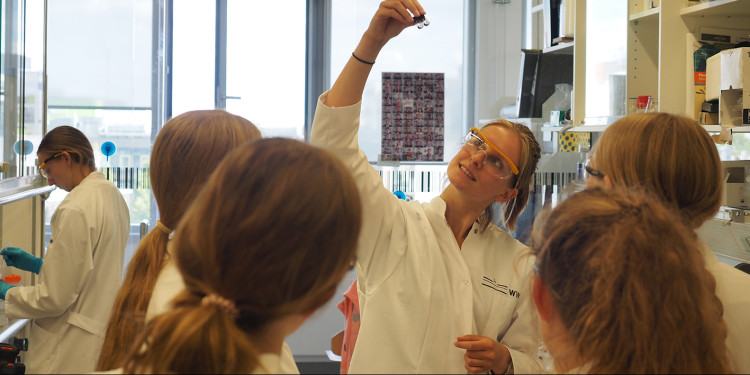 Schülerinnen im Labor, von hinten über die Schultern fotografiert. Den Schülerinnen zugewandt steht eine Kursleiterin. Sie hält ein kleines Probengefäß in der Hand und zeigt es den Schülerinnen. Alle abgebildeten Personen tragen weiße Laborkittel.<address>© MExLab Physik</address>