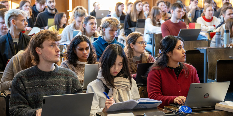 In einem Hörsaal sitzen zahlreiche Studierende. Einige von ihnen schreiben auf dem Laptop, andere schauen nach vorne auf den Dozenten oder die Präsentation.<address>© Uni MS - Johannes Wulf</address>