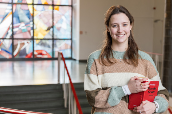 Die Jurastudentin Judith Boland steht in der Rechtswissenschaftlichen Fakultät, im Hintergrund ist eine bunte Glasfront zu sehen. Auf dem Arm trägt die angehende Juristin eine dickes, rotes Buch mit Gesetzestexten.<address>© Uni MS - Johannes Wulf</address>