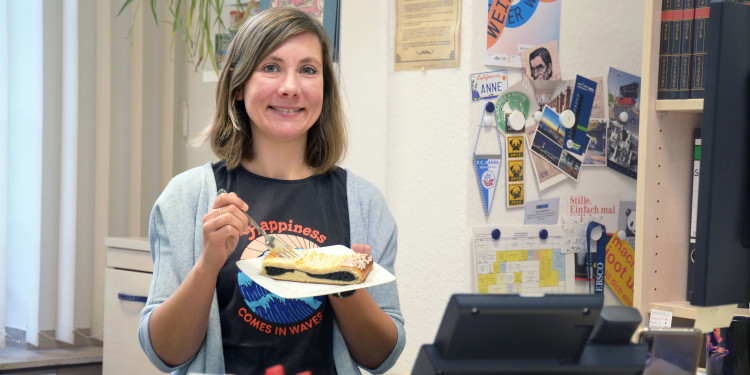 Eine Frau, Anne Sapich, steht in ihrem Büro hinter dem Schreibtisch und hält einen Teller mit einem Mohnkuchen in der Hand.<address>© Uni MS - Julia Harth</address>