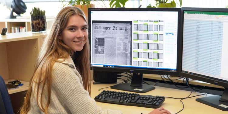 Eine junge Frau sitzt an einem Computerarbeitsplatz und schaut in die Kamera. Auf dem Computerbildschirm sind historische Zeitungsseiten zu sehen.<address>© Uni MS - Julia Harth</address>