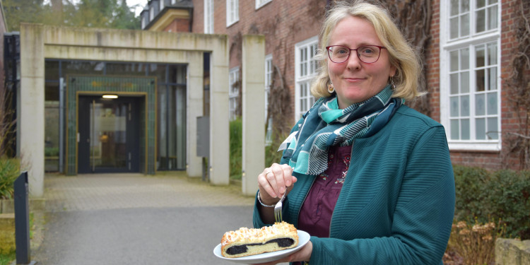 Monika Heitkötter steht vor dem Eingang zum Botanicum und schaut in die Kamera. In der Hand hat sie einen Teller mit einem Mohnkuchen.<address>&copy; Uni MS - Julia Harth</address>
