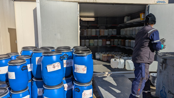 Auf dem Bild ist ein Containerbau zu sehen, dessen Türen geöffnet sind. Vor diesen stehen blaue Plastikfässer, rechts daneben steht ein Entsorgungsmitarbeiter. Im Container befinden sich Regale, in denen Kanister mit Chemikalien stehen.<address>&copy; Uni MS - André Bednarz</address>