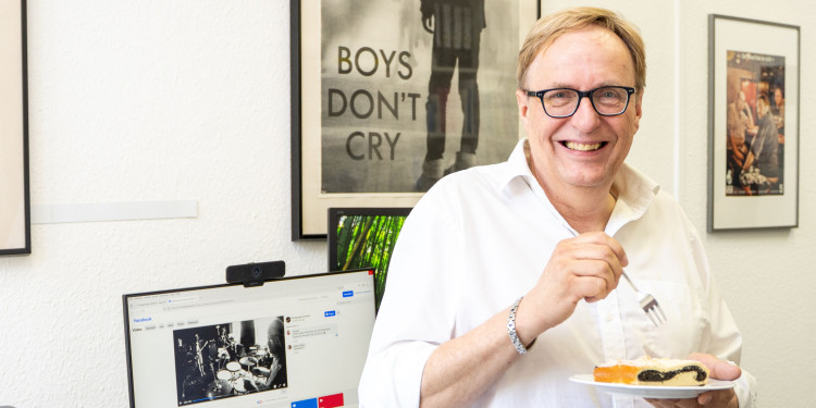 Das Bild zeigt Thomas Bauer, er lacht in die Kamera. In der rechten Hand hält eine Gabel, in der linken einen Teller mit einem Stück Mohnkuchen darauf. Im Hintergrund sind sein Schreibtisch mit einem Monitor sowie Konzertplakate zu sehen.<address>&copy; Uni MS - Johannes Wulf</address>