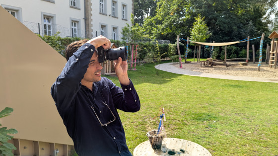 Das Bild zeigt Johannes Wulf mit einer Fotokamera. Er schaut durch dessen Sucher. Im Hintergrund ist ein Spielplatz der Unikita zu sehen.<address>© Uni MS - André Bednarz</address>
