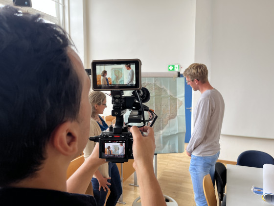 Auf dem Bild ist Johannes Wulf von hinten zu sehen. Er hält eine Filmkamera in der Hand. Im Hintergrund sind Karen Siegel und ein weiterer Mitarbeiter vom Institut für Politikwissenschaft zu sehen.<address>© Uni MS - André Bednarz</address>