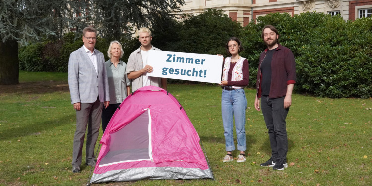 Mehrere Personen stehen hinter einem pinken Zelt und halten ein Schild mit der Aufschrift „Zimmer gesucht“ hoch. Im Hintergrund ist das Schloss zu sehen.<address>© Uni MS - Brigitte Heeke</address>