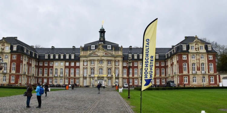 Zu sehen ist das Schloss in Münster. Vor dem Schloss steht eine sogenannte Beachflag mit dem Logo des Hochschultags.<address>© Uni MS - Linus Peikenkamp</address>