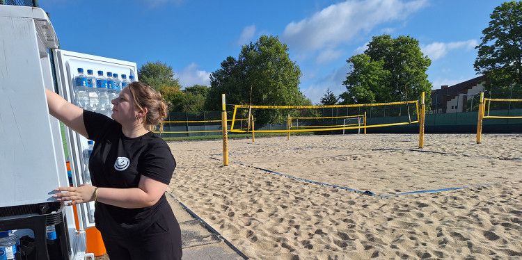Blick auf das Beachvolleyballfeld des Hochschulsports. Am linken Rand des Spielfelds steht Lara Barlag an einem geöffneten Kühlschrank. Sie blickt hinein und greift in den Kühlschrank.<address>© Uni MS - Christina Hoppenbrock</address>
