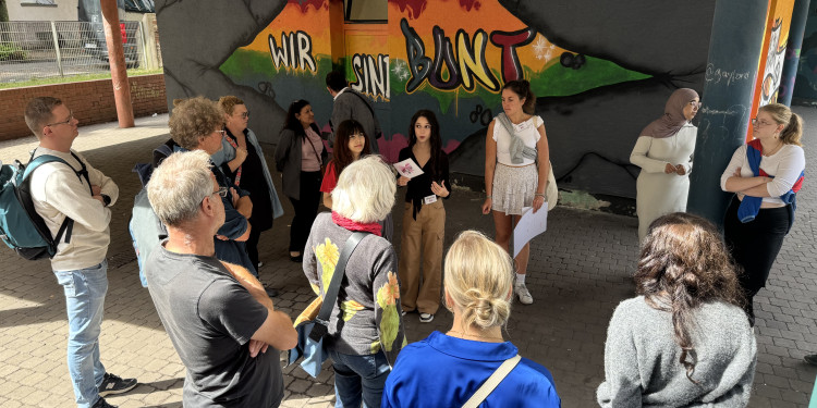 Das Bild zeigt eine Gruppe von rund 15 Personen. In der Mitte stehen die Schülerinnen Angelina und Esila, im Hintergrund steht auf einer Wand „Wir sind bunt“.<address>© Uni MS - Alice Büsch</address>
