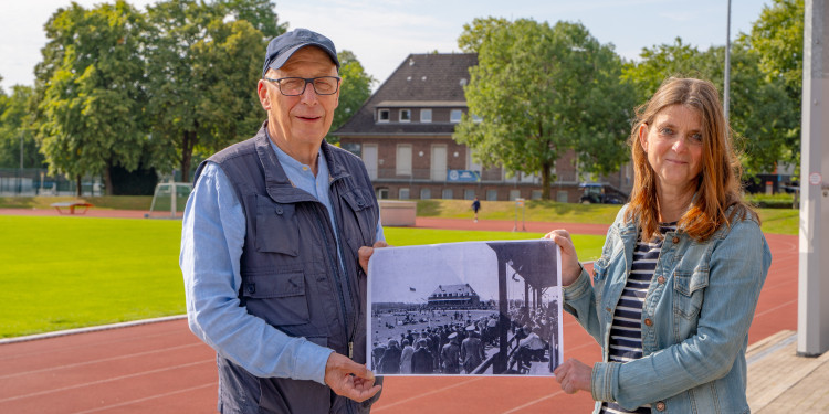 Das Bild zeigt Doktor Christiane Bohn und Professor Doktor Dieter Heinz Jütting. Sie stehen vor einem Sportplatz. Sie halten ein Foto desselben Ortes von 1929 in der Hand. Zu dieser Zeit fanden die Deutschen Hochschulmeisterschaften statt.<address>© Uni MS - Linus Peikenkamp</address>
