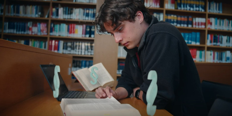 Das Foto zeigt einen Studenten. Er sitzt in einer Blbliothek und liest ein Buch. Vor ihm stehen animierte Paragraphen.<address>© Malte Papenfuss</address>