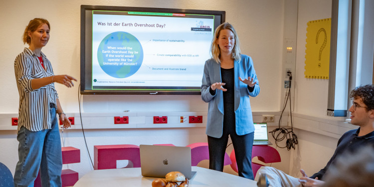 Die Seminarleiterinnen Ronja Dobler und Dr. Lea Püchel (v. l.) erläutern den Studierenden, was der Earth Overshoot Day ist.<address>© Uni MS - Linus Peikenkamp</address>