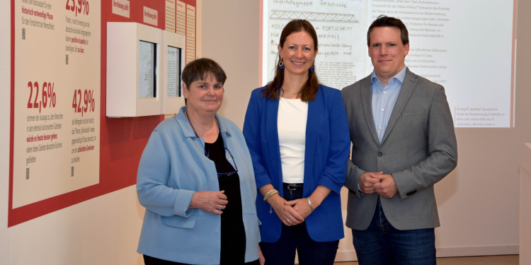 Das Bild zeigt zwei Frauen und einen Mann, die in der Ausstellung im Stadtmuseum stehen und in die Kamera schauen. Links an der Wand sind Statistik-Zahlen erkennbar, im Hintergrund eine Beamer-Präsentation mit Forschungsergebnissen.<address>© Uni MS - Julia Harth</address>