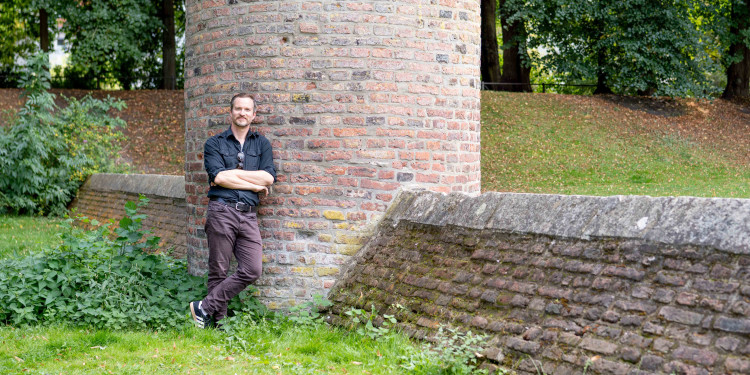 Dr. Jan Kortmann leht an einem kleinen gemauerten Turm mit Mauer an der Promenade in Münster auf einer Wiese.<address>© Uni MS - Linus Peikenkamp</address>