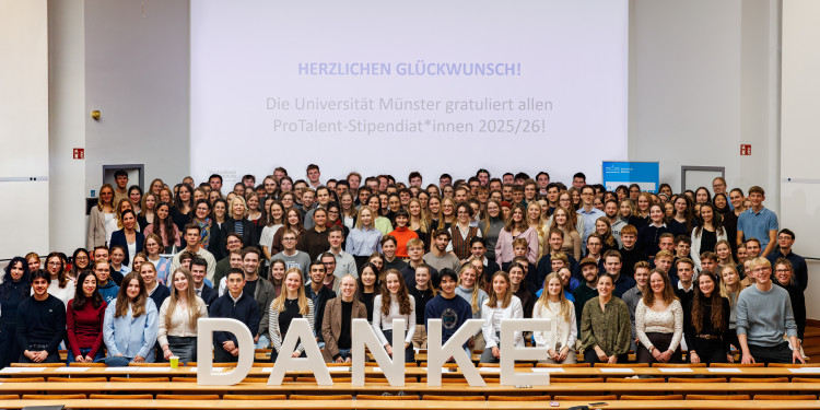 Die Studierenden, die durch das ProTalent-Programm unterstützt werden, stehen in einem Hörsaal, vor ihnen formen fünf große weiße Buchstaben das Wort &quot;DANKE&quot;. Alle lachen in dir Kamera.<address>© Uni MS - Peter Leßmann</address>