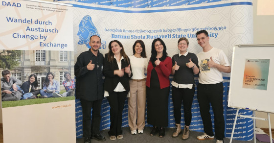 The picture shows six people smiling at the camera in front of posters and billboards advertising the Spring School in Georgia.<address>© University of Münster - German Studies Department</address>