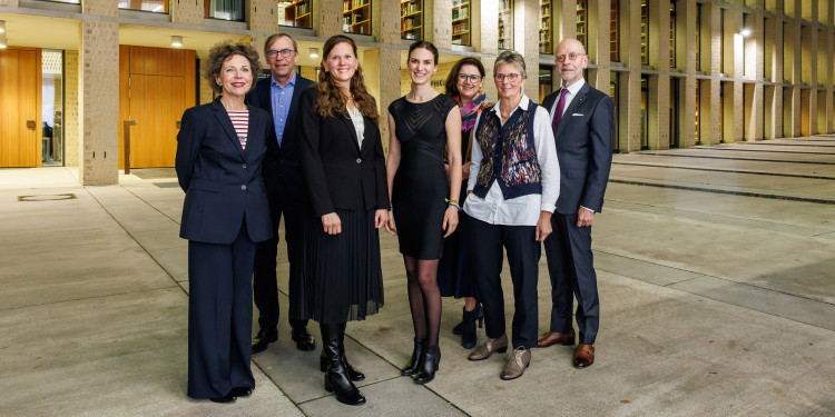 Sieben Personen, darunter Anna-Maria Balbach, Jana Haack und Barbara Schüler in der Bildmitte, stehen vor dem beleuchteten Philosophikum der Uni Münster und blicken in die Kamera.<address>© Uni MS - Peter Leßmann</address>