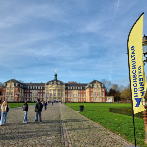 Zu sehen ist das Schloss Münster, mit einer Fahne des Hochschultags im Vordergrund und einigen jungen Menschen, die in Richtung Schloss gehen.<address>© Uni MS - Brigitte Heeke</address>