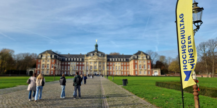 Zu sehen ist das Schloss Münster, mit einer Fahne des Hochschultags im Vordergrund und einigen jungen Menschen, die in Richtung Schloss gehen.<address>© Uni MS - Brigitte Heeke</address>