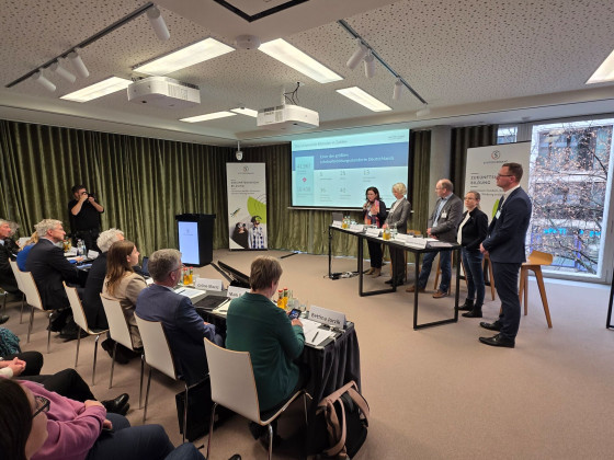 Die Aufnahme entstand während der Präsentation des Teams der Universität Münster. Alle Personen stehen hinter einem Tisch und haben Unterlagen vor sich liegen. An der Wand ist die Präsentation zu sehen.<address>© Uni MS - Norbert Robers</address>