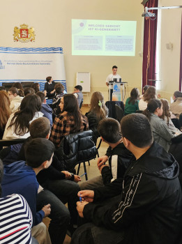 The picture shows participants in a room listening to a young man at the lectern. A presentation can be seen in the background.<address>© University of Münster - German Studies Department</address>