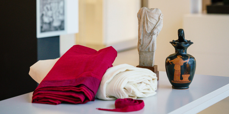 Zu sehen ist eine Statuette aus dem antiken Griechenland neben einem kleinen verzierten Tongefäß sowie zwei Rollen Stoff in den Farben rot und weiß.<address>© Archäologisches Museum Münster</address>