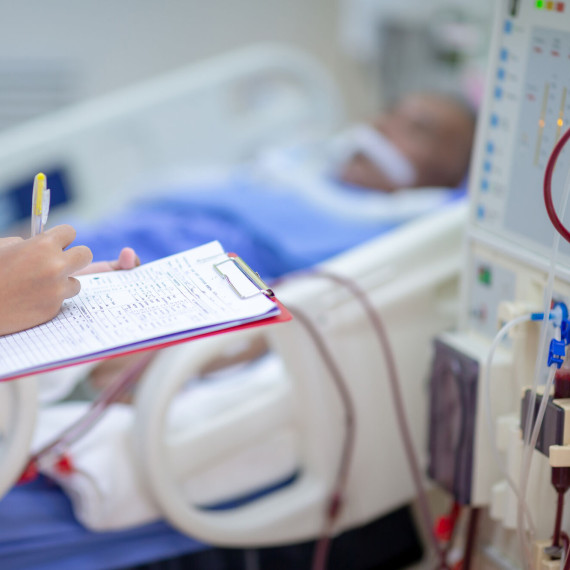 Medizinische Fachkraft hält ein Klemmbrett und dokumentiert Werte, während neben einem Krankenhausbett eine Dialysemaschine mit Schläuchen und blutgefülltem Filter zu sehen ist; der Patient liegt unscharf im Hintergrund.<address>© stock.adobe.com - Hospital man</address>