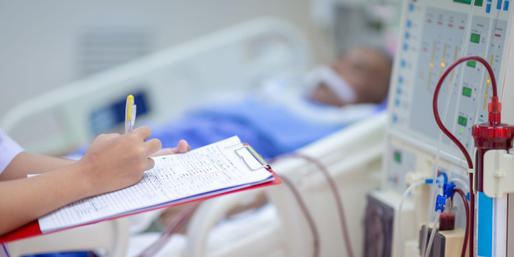 Medizinische Fachkraft hält ein Klemmbrett und dokumentiert Werte, während neben einem Krankenhausbett eine Dialysemaschine mit Schläuchen und blutgefülltem Filter zu sehen ist; der Patient liegt unscharf im Hintergrund.<address>© stock.adobe.com - Hospital man</address>