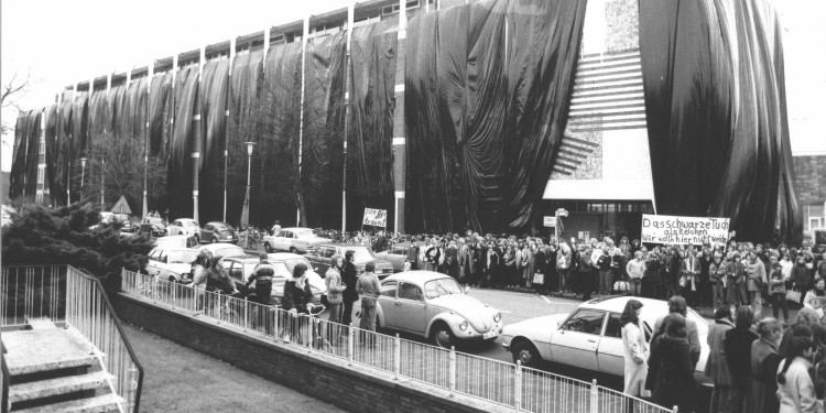 Die Schwarz-weiß-Aufnahme zeigt das Zentralgebäude der Pädagogischen Hochschule an der Scharnhorststraße, das in Anlehnung an Aktionen des Verpackungskünstlers Christo mit Plastikplanen verhüllt ist. Im Vordergrund stehen viele geparkte Autos am Straßenrand. Außerdem sind Dutzende Menschen auf den Gehwegen zu sehen, die zum Teil Plakate in die Luft halten.<address>&copy; Norbert Breuer, Warendorf</address>