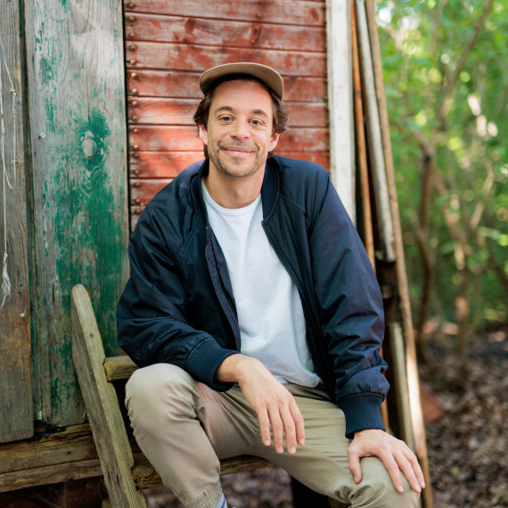 Das Bild zeigt Tobias Krell mit weißem Shirt, dunkelblauer Jacke und Schirmmütze. Er sitzt auf einer Holzleiter vor einer Holzhütte im Wald und schaut lächelnd in die Kamera.<address>&copy; Hans-Florian Hopfner</address>