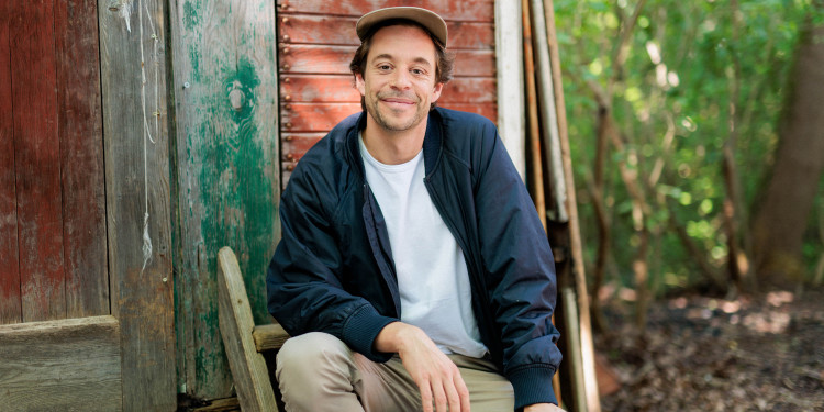 Das Bild zeigt Tobias Krell mit weißem Shirt, dunkelblauer Jacke und Schirmmütze. Er sitzt auf einer Holzleiter vor einer Holzhütte im Wald und schaut lächelnd in die Kamera.<address>&copy; Hans-Florian Hopfner</address>