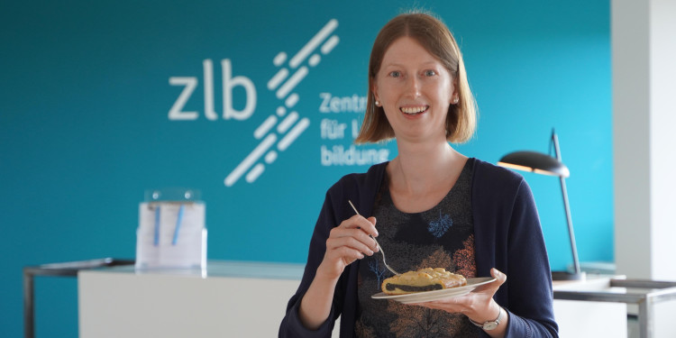 Das Bild zeigt Anna-Frederike Gunkler mit einem Mohnkuchen auf einem Teller in der Hand im Eingangsbereich des ZLB. Im Hintergrund an der blauen Wand ist der Schriftzug des ZLB erkennbar.<address>&copy; Uni MS - Julia Harth</address>