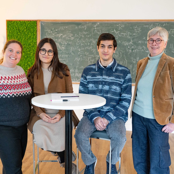 Das Bild zeigt vier Personen, die hinter einem Stehtisch sitzen und stehen und in die Kamera schauen. Im Hintergrund ist eine grüne Tafel mit Matheformeln zu sehen.<address>&copy; Uni MS - Victoria Liesche</address>