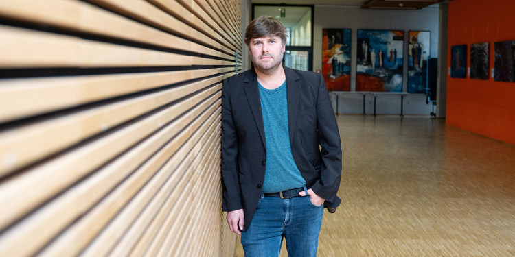 Physiker Prof. Dr. Tobias Heindel im Foyer des Center for Nanotechnology (CeNTech). Er ist an eine Wand gelehnt, die mit horizontalen Holzlatten verkleidet ist. Tobias Heindel steht mittig im Bild, einige Meter entfernt vom Fotografen. Die linke Bildhälfte ist von der Wand ausgefüllt, rechts im Hintergrund ist das Foyer zu sehen.<address>© Uni MS - Linus Peikenkamp</address>