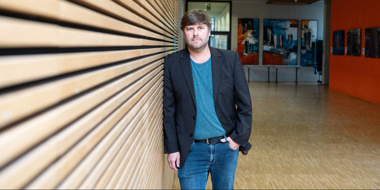 Physicist Prof. Dr. Tobias Heindel in the foyer of the Centre for Nanotechnology (CeNTech). He is leaning against a wall covered with horizontal wooden slats. Tobias Heindel stands in the centre of the picture, a few metres away from the photographer. The left half of the picture is filled by the wall, while the foyer can be seen in the background on the right.<address>© Uni MS - Linus Peikenkamp</address>