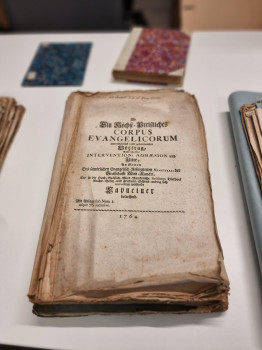 Zu sehen ist im Vordergrund eine Beispielseite aus dem Konvolut, einem Buchtitel. Im Hintergrund sind weitere Archivalien zu sehen.<address>© Uni MS - Brigitte Heeke</address>