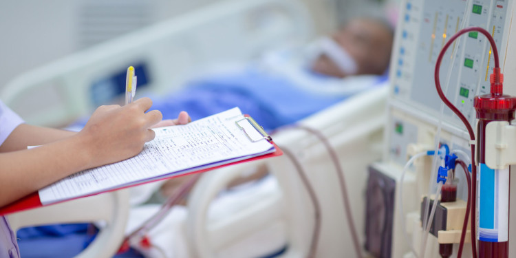 A healthcare professional holds a clipboard and records values while a dialysis machine with tubing and a blood-filled filter stands beside a hospital bed; the patient is blurred in the background.<address>© stock.adobe.com - Hospital man</address>