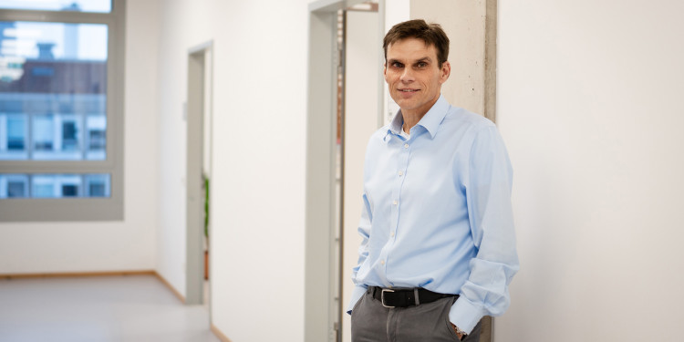 Prof. Dr. Armido Studer im Porträt. Er trägt ein hellblaues Hemd und eine dunkelgraue Hose und steht in einem Flur mit weißer Wand, den Rücken nach rechts in Richtung Wand gerichtet, dem Betrachter halb zugewandt. Die Flurflucht läuft nach links auf ein Fenster zu.<address>© Uni MS - Linus Peikenkamp</address>