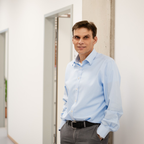 Portrait of Prof. Armido Studer. He is wearing a light blue shirt and dark grey trousers and is standing in a hallway with white walls, his back to the right towards the wall, half-facing the viewer. The hallway runs to the left towards a window.<address>© Uni MS - Linus Peikenkamp</address>