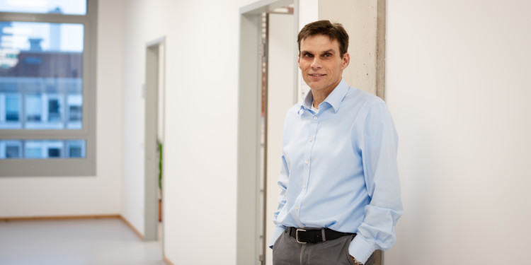 Portrait of Prof. Armido Studer. He is wearing a light blue shirt and dark grey trousers and is standing in a hallway with white walls, his back to the right towards the wall, half-facing the viewer. The hallway runs to the left towards a window.<address>© Uni MS - Linus Peikenkamp</address>