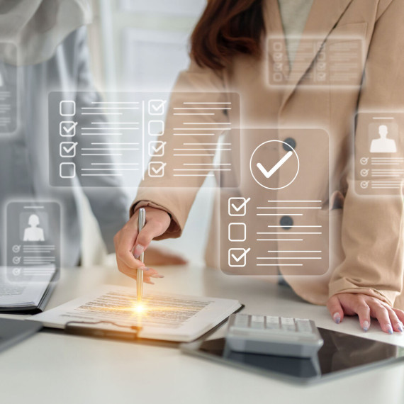 Zwei Personen stehen an einem Schreibtisch in einem Büro. Eine Person markiert mit einem Stift ein Dokument auf einem Klemmbrett. Auf dem Tisch liegen ein Laptop, ein Tablet und ein Taschenrechner. Über dem Bild sind symbolisch digitale Checklisten und Häkchen eingeblendet, die für Prüfung, Auswahl oder Bewertung stehen.<address>&copy; stock.adobe.com - STBSTD</address>